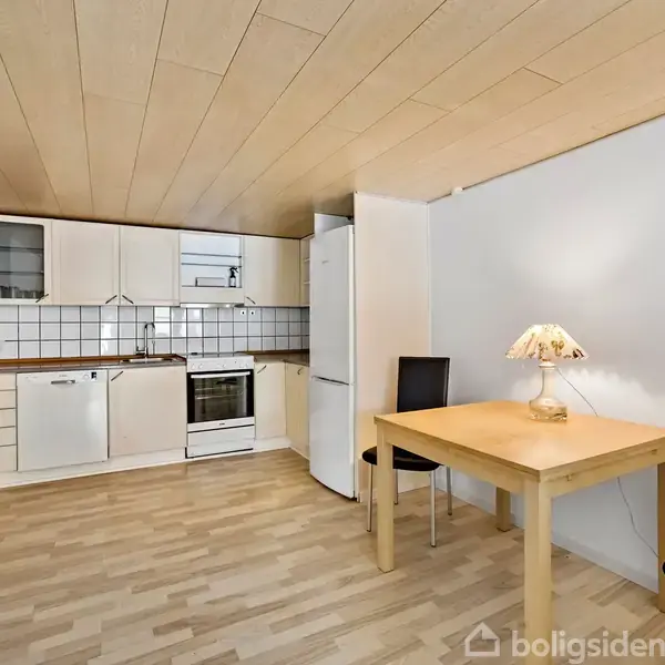 Et køkken har lyse træskabe og apparater, inklusive en ovn og opvaskemaskine. Spisebordet med to stole står i et hjørne, belyst af en bordlampe. Laminatgulv og træloft kompletterer rummet.