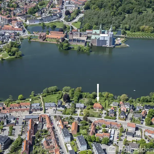 En by set fra oven, hvor hovedfokus er en sø omgivet af træer og bygninger. En markør viser en specifik ejendom nær søen. I baggrunden ses et stort slot.