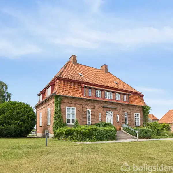 Et rødt murstenshus med rødt tegltag står omgivet af en velholdt græsplæne og buske. Huset har hvide vinduer og en trappe ved hovedindgangen, beliggende i et grønt område.