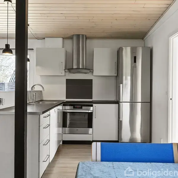 Et moderne køkken med hvide skabe og stålinventar; køleskab og ovn integreret i højre side. Spisebordsområde med en blå bænk i forgrunden; vinduer i baggrunden slipper lys ind.