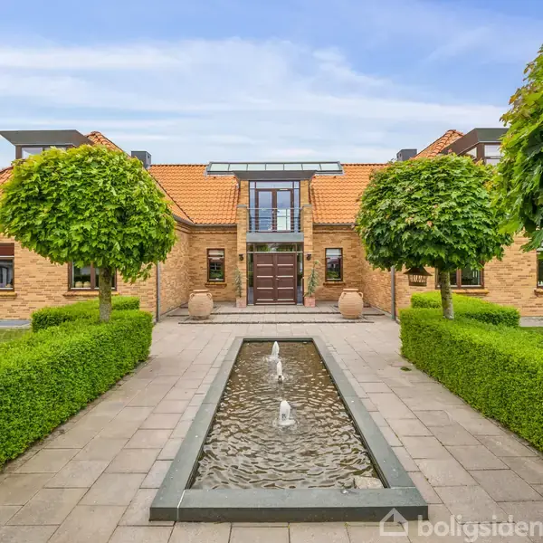 En stor murstensvilla med rødt tegltag står i centrum. Foran er en lang rektangulær springvand omgivet af flisebelagte stier og hæklabyrinter med træer.