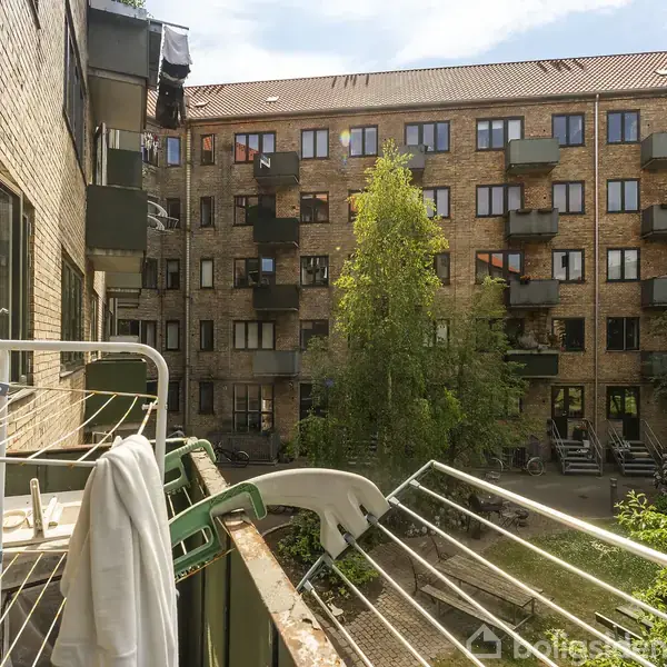 En altan med tørrestativ og håndklæde står mod en gård omgivet af en murstensbygning med flere altaner. En lille have med træer ses i gården.