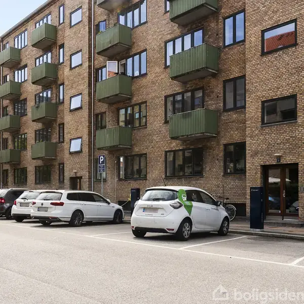 En murstensbygning med grønne altaner står langs en gade. Flere biler er parkeret foran bygningen. En parkeringsskilt ses ved siden af bilerne.
