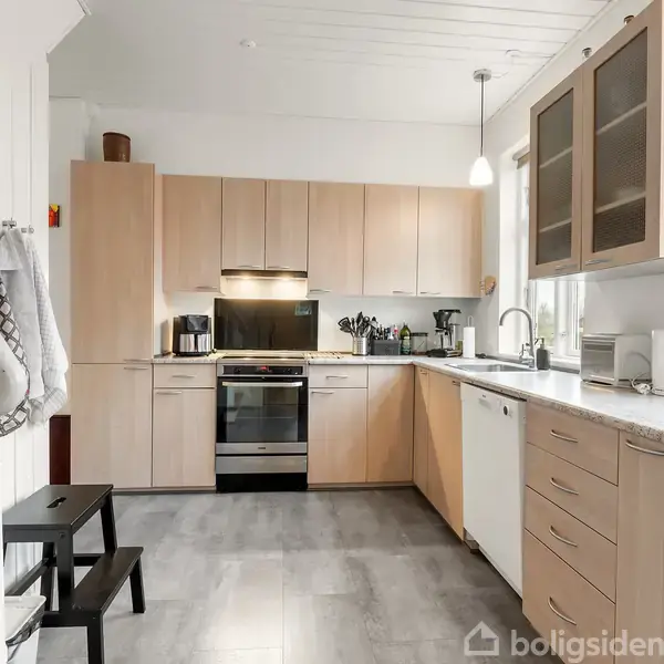 Køkken med lyse træskabe opstillet i en vinkel, moderne komfur i midten. Grå flisegulv, opvaskemaskine til højre, hvid vask. Overbelysning og vindue mod højre, trappe til venstre.