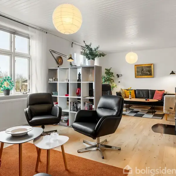 To sorte lænestole står ved et rundt sofabord i en lys stue med trægulv. Bagved er en reol med pynteting og planter, mens en sofa og brændeovn ses i baggrunden.