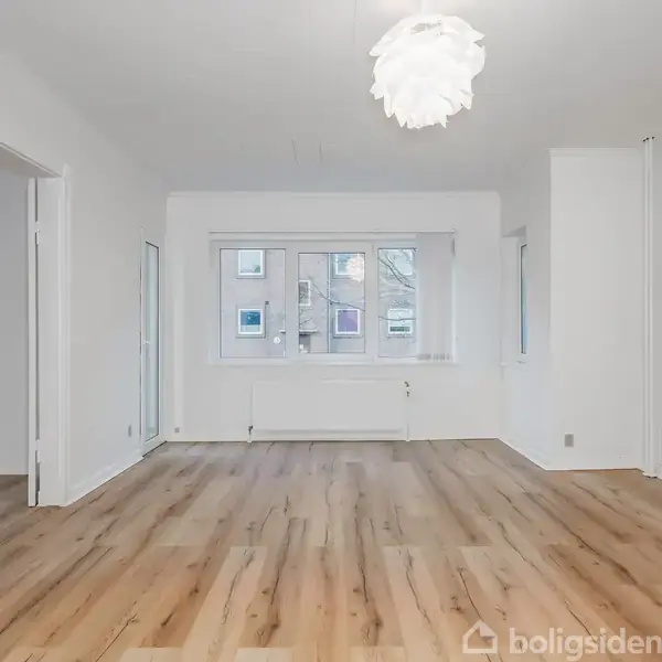Et lyst rum med trægulve; en moderne, blomsterformet loftslampe hænger ned. Rummet indeholder vindue med udsigt til bygninger udenfor, og en radiator befinder sig under vinduet.