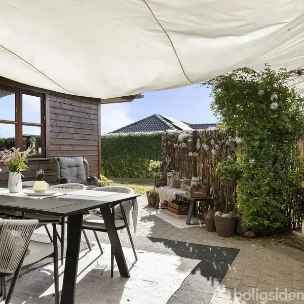 Terrasse med spisebord og stole, overdækket af stort solsejl. I baggrunden ses et træhus, grønne planter og en udendørs kamin. Sollys skinner gennem sejl.