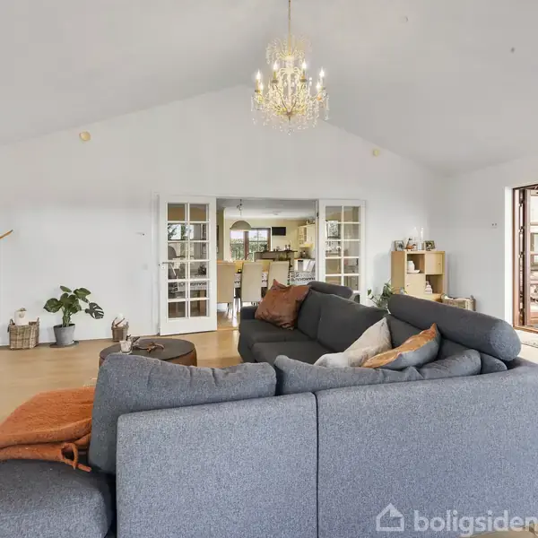 En stor grå sofa står centralt i en lys stue. Rummet er dekoreret med planter, en lysekrone, og åbner op mod en spisestue med et langbord og flere stole.