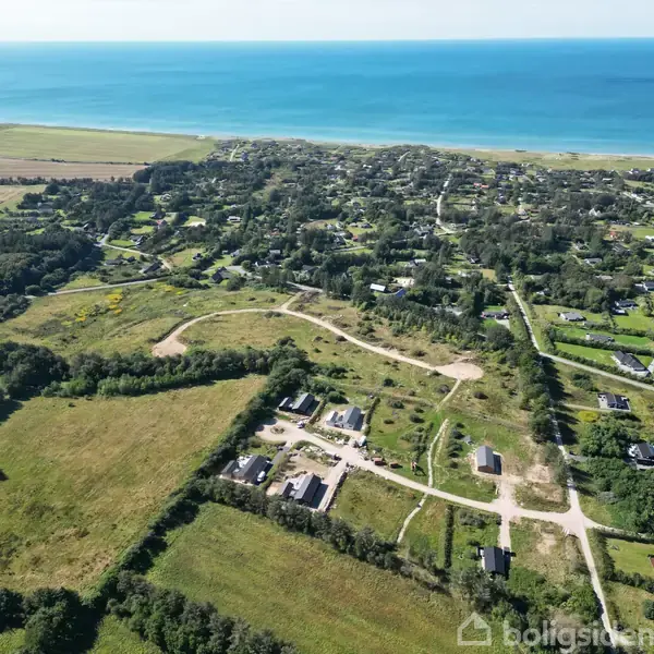 Luftfoto viser et landskab med spredte huse og veje, omgivet af grønne marker og træer. I baggrunden ses havet, der strækker sig ud i horisonten.