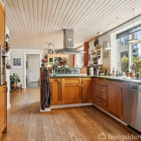 Et moderne køkken med træskabe og gulve. En stål-emhætte hænger over kogepladen. Store vinduer på højre side lader sollys ind, og der er planter i baggrunden.