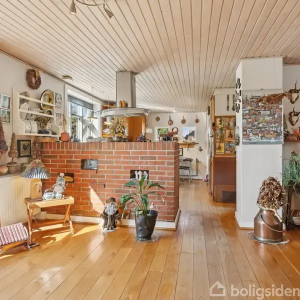 Stue med trægulv og loft, indeholder et åbent køkken med en skillevæg af mursten. Dekoreret med planter, malerier og jagttrofæer, og sollys strømmer ind gennem store vinduer med røde gardiner.