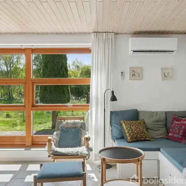 Stue med blå sofa og puder ved siden af et rundt sofabord og en lænestol. Store vinduer med udsigt til grøn natur, træbord til spisning, hvidmalet væg med kunst.