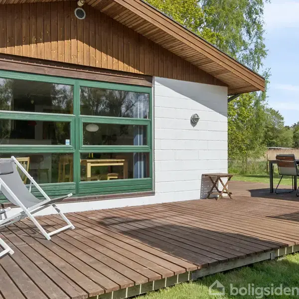 Træterrasse med to liggestole foran et hvidt hus med store vinduer. Rundt om står et bord med stole og en parasol. Græsplæne og træer i baggrunden.
