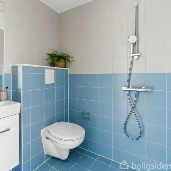 Et badeværelse med blå fliser har en væghængt toilet og en bruser. Ved håndvasken står sæbedispensere og tandbørstekrus. En plante står ovenpå en lille hylde.