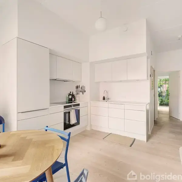 Et rundt spisebord med blå stole står i lyse omgivelser foran store vinduer, der fører ud til en balkon. Der er planter og en rød bygning i baggrunden.
