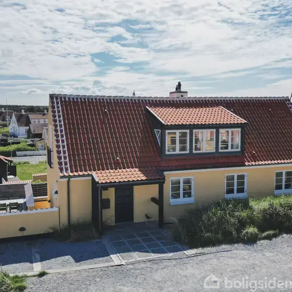 Et gult hus med rødt tegltag står stille på en grusvej. I baggrunden ses en række andre huse i et boligområde under en delvist skyet himmel.