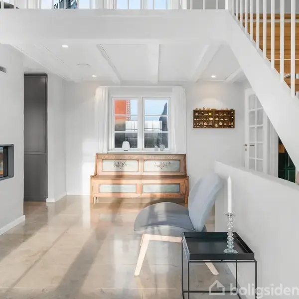 Skulpturelt trappeopgang leder til overetage i moderne stue. Indbygningspejs til venstre, glasbord med stearinlys til højre. Sollys strømmer ind gennem vindue over en lave reol.