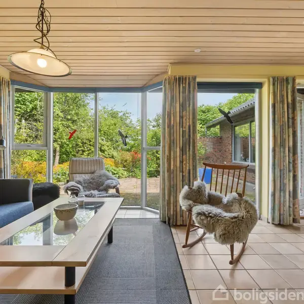 En stue med en blå sofa og glasbord foran et stort vindue. Lænestol med pels og gyngestol står ved siden af. Udsigt til en grøn have.