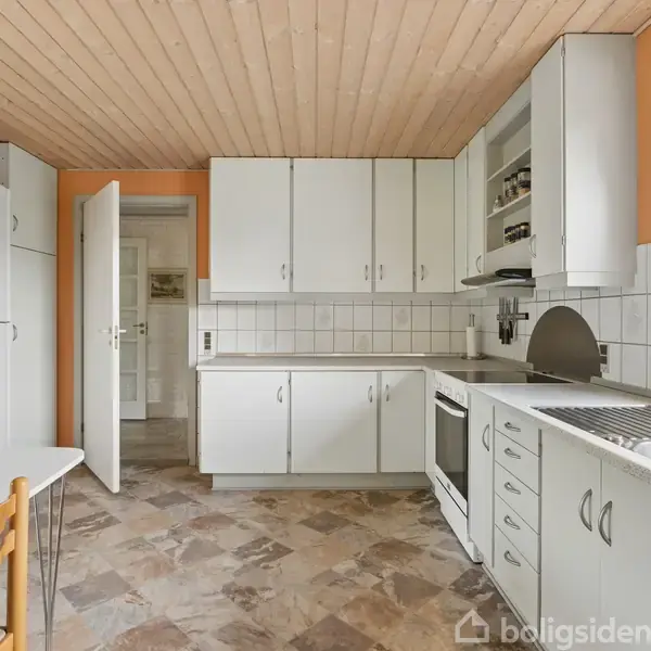 Køkken med hvide skabe og skuffer, en indbygget ovn, håndvask samt et lille bord med to stole. Gulvet er mønstret, og loftet er af træ. En dør står åben i baggrunden.