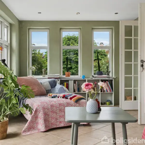 Plante står i en kurve ved siden af en sofa dækket med et tæppe, mens en lyserød blomst på et bord skaber hygge. Store vinduer viser grøn udsigt.