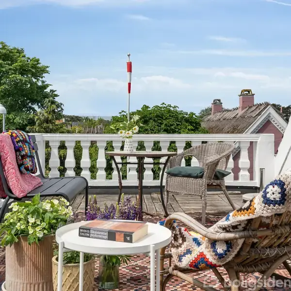 Balkon med møblerede stole og et bord viser tæpper og blomsterdekorationer. En bog ligger på bordet ved siden af en plante. Udsigt til træer og huse i baggrunden.