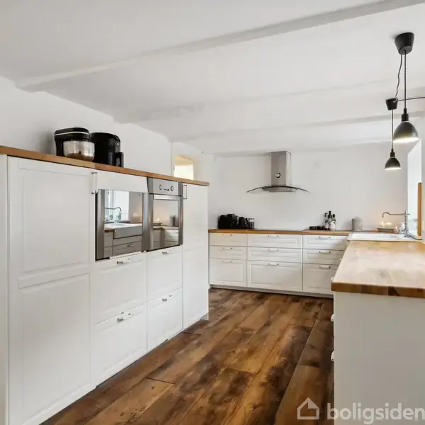 Hvidt køkken med træborde og moderne apparater, herunder indbyggede ovne. Trægulv strækker sig gennem rummet, som oplyses af hængende lamper. Enkel dekoration inkluderer planter på vindueskarmen.