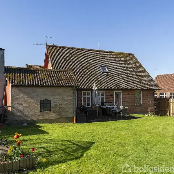 Hus med sadeltag og murstensvægge ligger i en grøn have. Til højre ses et hegn, og til venstre står et drivhus. Terrasse med havemøbler ved siden af huset.