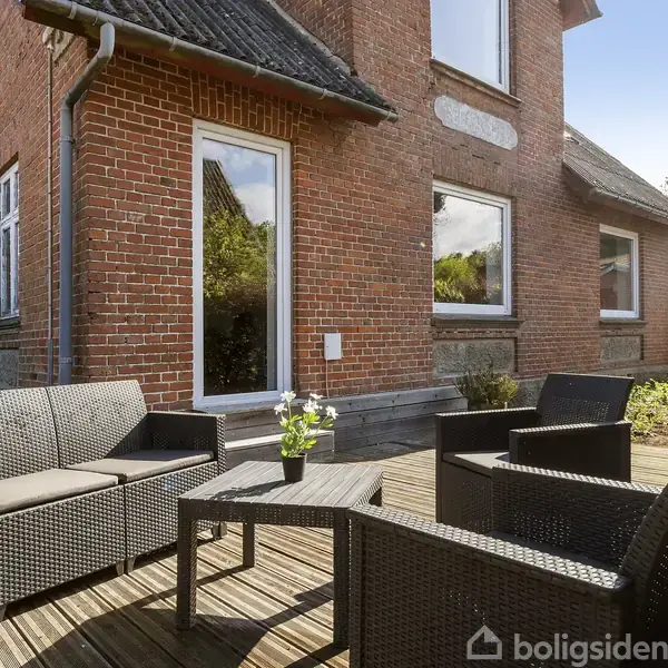 Terrassemøbler står på en træterrasse foran et rødstenshus. Fire kurvestole og et bord er arrangeret omkring en plante på bordet, omgivet af grønne buske og et hegn.