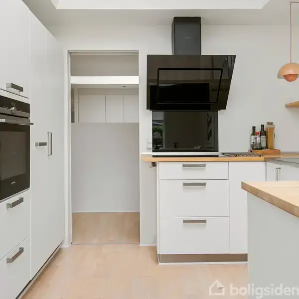 Et moderne køkken med hvide skabe, integrerede ovne og en sort emhætte. Bordplader i træ omgiver vasken og kogepladen. En orange lampe hænger over bordet.