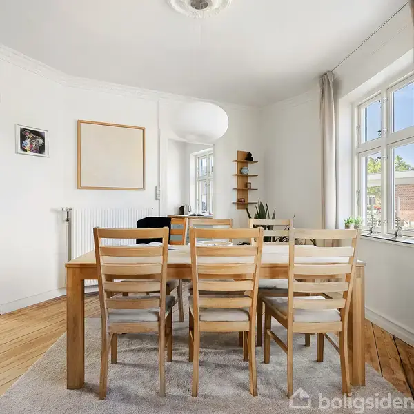 Spisebord med otte stole står i et lyst rum med trægulv. På væggene hænger billeder, og der er vinduer med hvide gardiner. En hylde og planter fylder hjørnet.