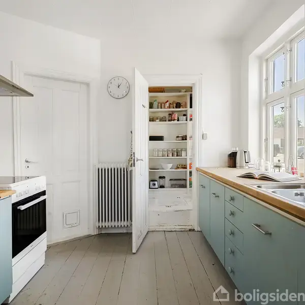 Køkken med blå skabe, åben skabslåge viser hylder fyldt med madvarer. Komfur og vask er ved siden af hinanden. Vægur hænger på væggen imellem døre og vindue.
