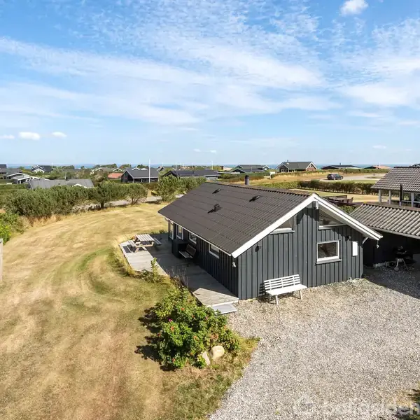 Et sort træhus står stille omgivet af en grusbelagt indkørsel og en tør græsplæne. I baggrunden ses flere huse og en blå himmel.