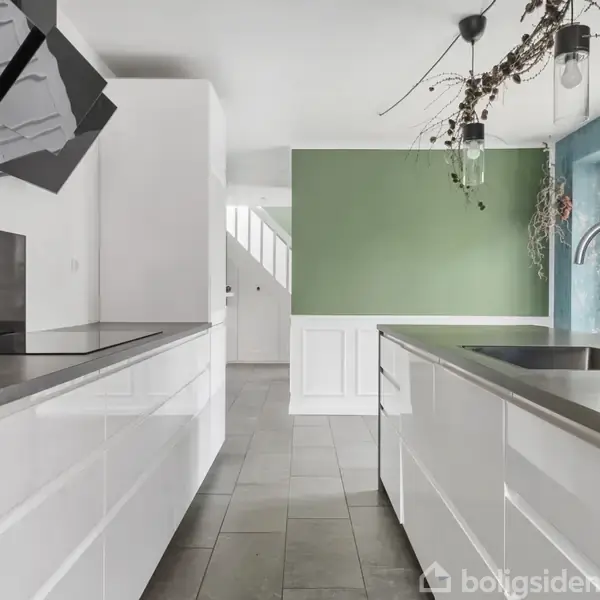 Et moderne køkken med hvide skabe og grå bordplader, hvor en stor emhætte er monteret. Spisebord med grene hænger fra loftet. Væggene er grønne med et vindue til højre.