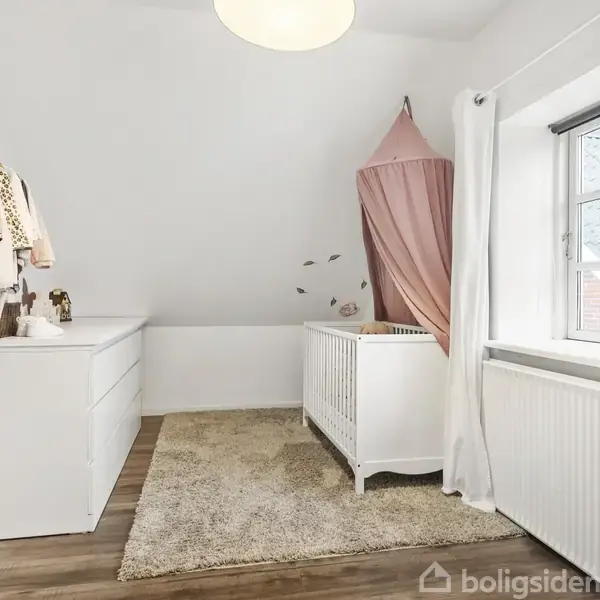 Værelse med en hvid tremmeseng under en lyserød baldakin. Til venstre hænger tøj fra en bøjlestang over en kommode. Vindue til højre med hvidt gardin og radiator.