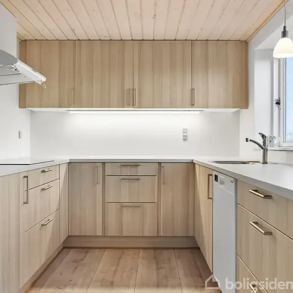 Et moderne køkken med lyse træskabe, integreret ovn og emhætte. Skabene er arrangeret i en L-form, og der er en hvid bordplade. Et vindue slipper lys ind i rummet.