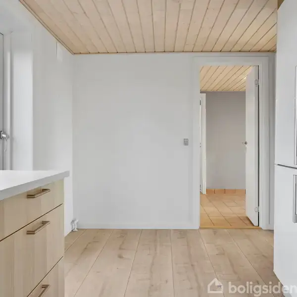 Et moderne køkken med lyse træmøbler og hvide vægge. En åben dør fører til en gang. Lyse trægulve og loft tilføjer et naturligt præg.