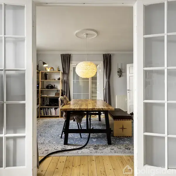 Et bord står i et rum med lyse vægge og trægulv. Store glasdøre åbner til et rejst område med en stor lampe og reol. En fjernsynsskærm ses til venstre.