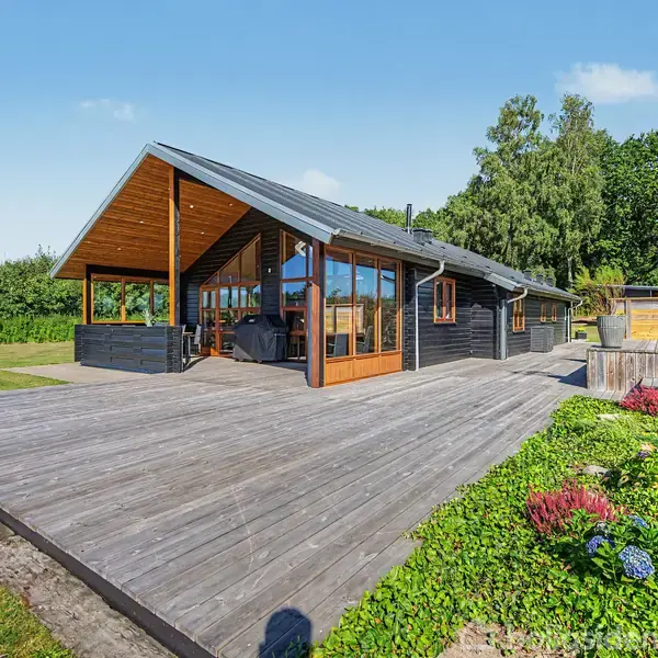 Sort træhus med store vinduer står på en bred træterrasse. Huset er omgivet af grønne buske og græs, med en blå himmel ovenfor.