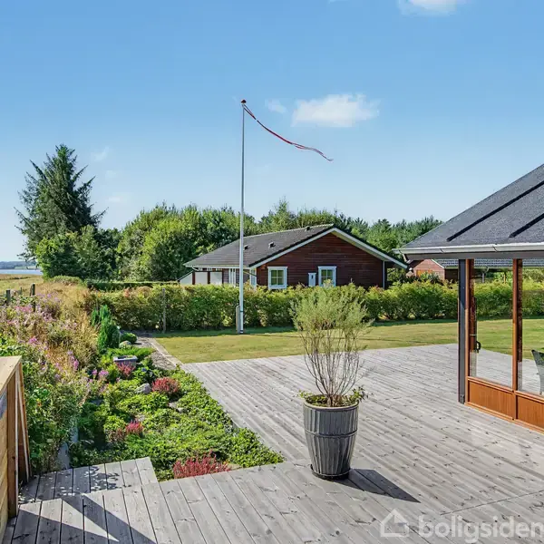 En træterrasse med en plantekrukke foran et moderne hus. Bagved er en stor græsplæne, en rødklædt bygning og en flagstang. Landskabet består af grønne marker og skove.