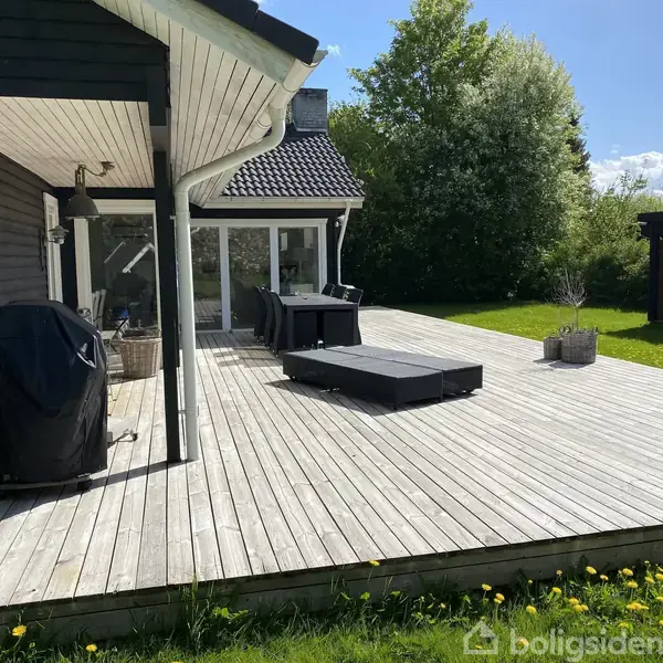 En stor træterrasse med liggestole og et spisebord sætter scenen foran et moderne hus. Haven er grøn og omgivet af træer under en blå himmel med skyer.