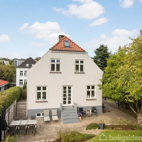 Hus med rødt tegltag, hvid facade og tre etager. Foran huset er en terrasse med havemøbler, omkranset af have med træer. Andre huse er synlige i baggrunden.