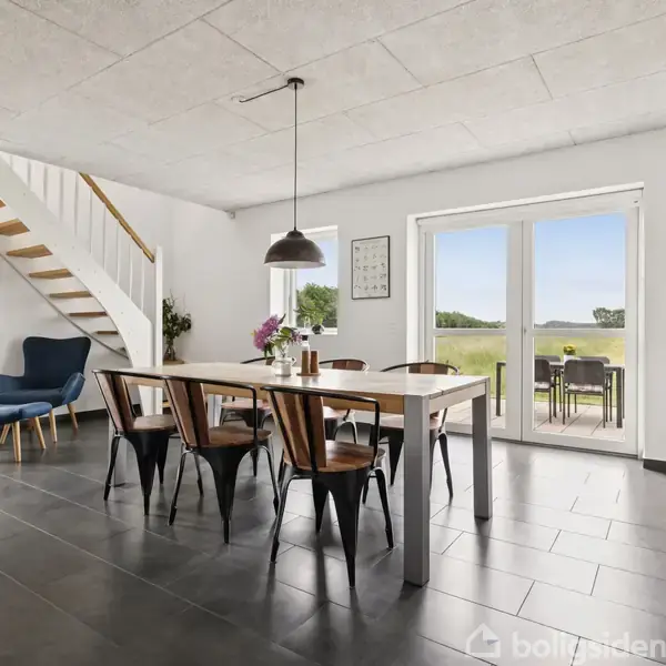 Et moderne spisestueområde med et træbord og seks stole, placeret under en hængelampe. Trappen fører opad, og store glasdøre åbner til en terrasse med udsigt over en mark.
