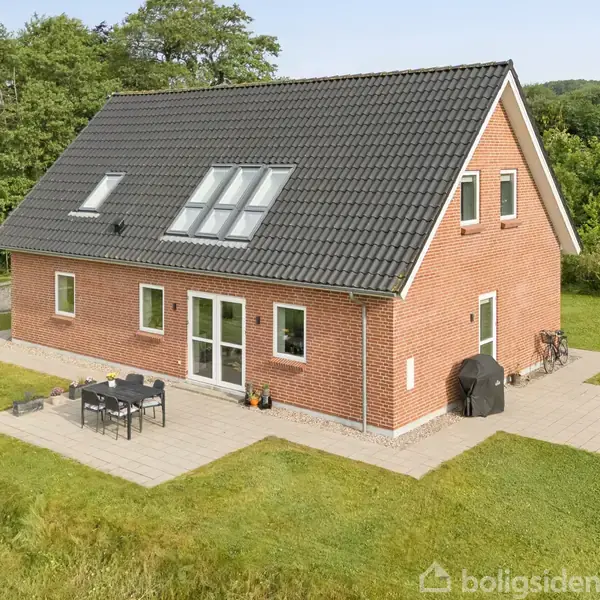 Et rødt murstenshus står på en græsplæne; huset har sort tag, flere vinduer og en terrasse med havemøbler. Det er omkranset af buske og træer.