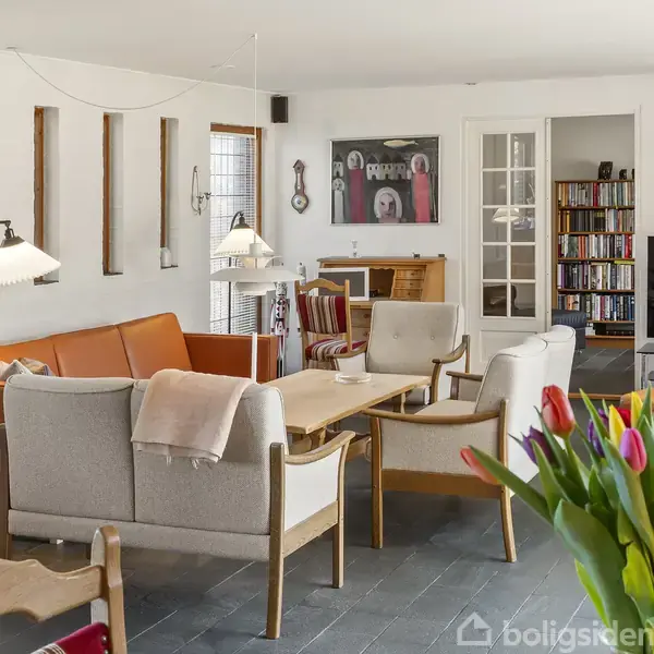 Stue med møbler: En komfortabel sofa og stole omkring et sofabord, omgivet af reoler og kunst på væggene. Tulipaner i en vase på bordet tilføjer farve til rummet.