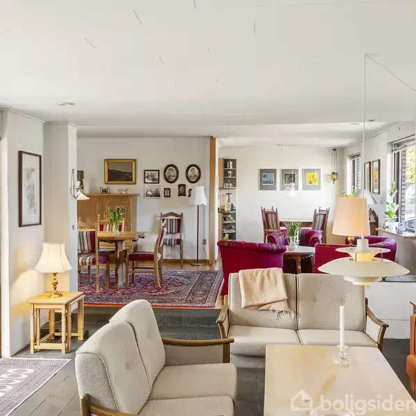 Stue med lyse sofaer og et kaffebord i midten, omgivet af farverige malerier på væggen. I baggrunden ses spisebord med stole og reoler. Store vinduer giver naturligt lys.