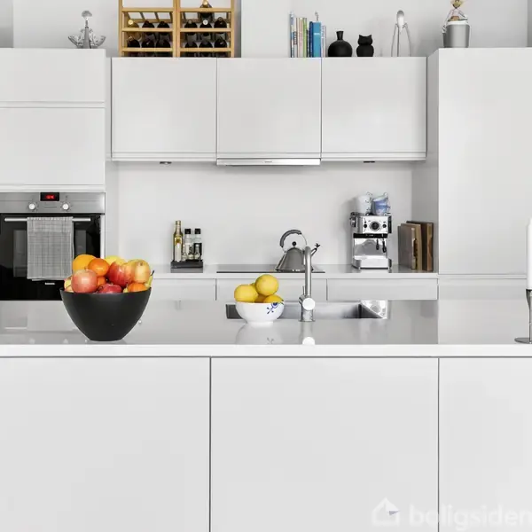 Hvidt køkken med moderne indretning. Frugtskåle med æbler og citroner står på køkkenbordet. En ovn og espressomaskine er integreret. Bøger og dekorationer pryder hylderne.