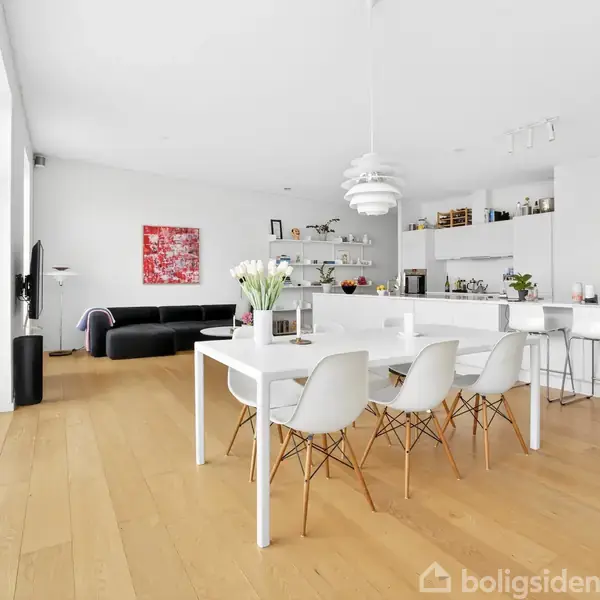 Stort, lyst køkken-alrum med hvidt spisebord og moderne stole. I baggrunden ses en sofagruppe og vægkunst. Rummet har trægulv, store vinduer og minimalistisk indretning.