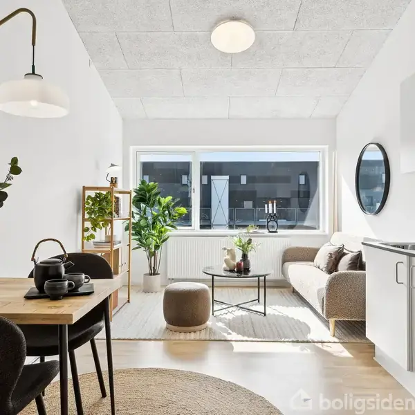 En moderne stue med en sofa, et rundt sofabord og en gulvplante. Til venstre er der et spisebord med to stole og en lampe. Store vinduer giver lys.