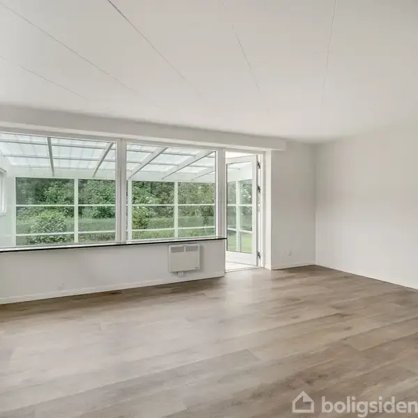 Stue med trægulv ligger stille, omgivet af lyse vægge og store vinduer, der åbner mod en overdækket terrasse med udsigt til grøn natur.