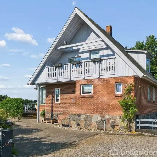 Et rødt murstenshus med en hvid balkon og skråt tag står i en gårdsplads med grus. Omgivet af grønne træer under en blå himmel med få hvide skyer.
